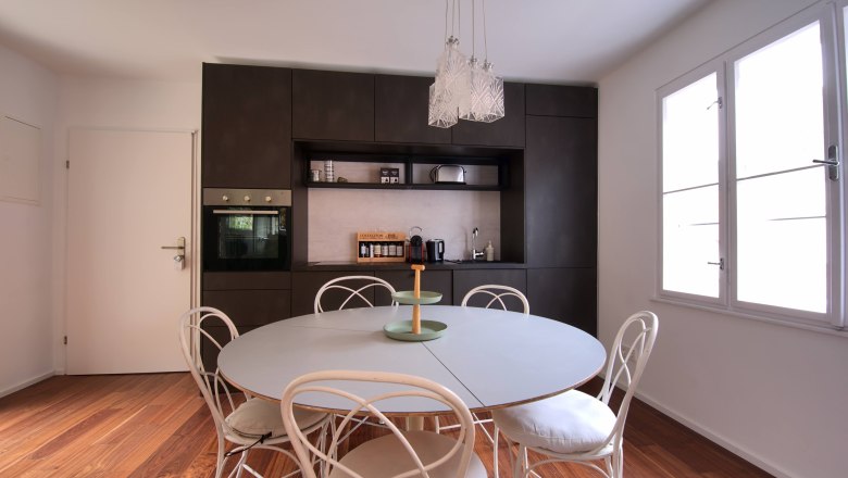 Modern dining area with round table, white chairs and dark kitchen unit in the background.