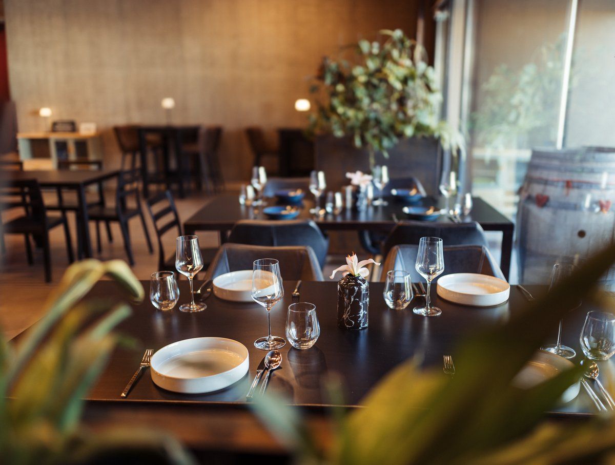 Elegant restaurant with laid tables, wine glasses and plants in the background.