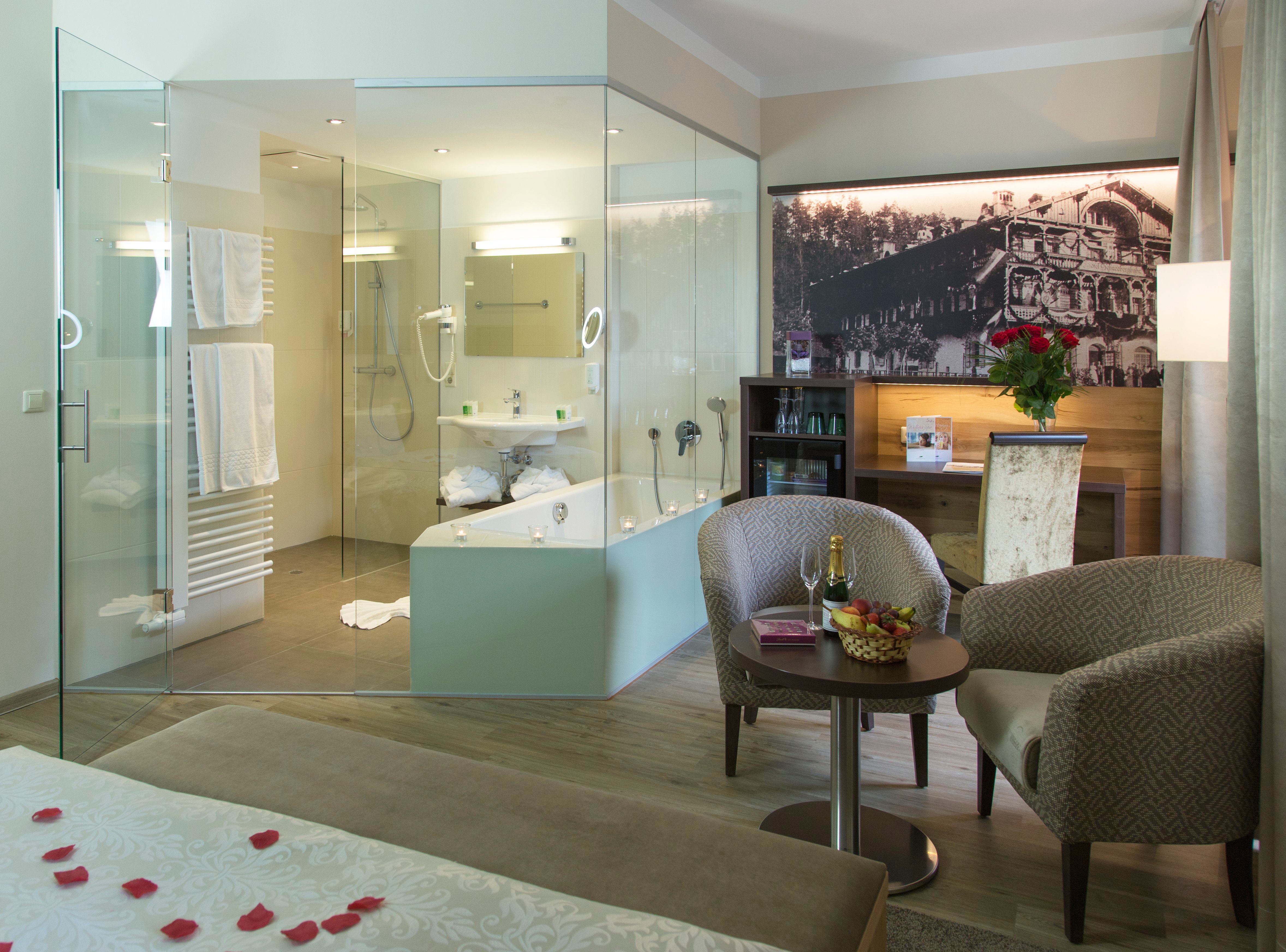 Luxurious hotel room with glass bath, cozy armchairs, table with champagne and fruit, and a large bed with rose petals.
