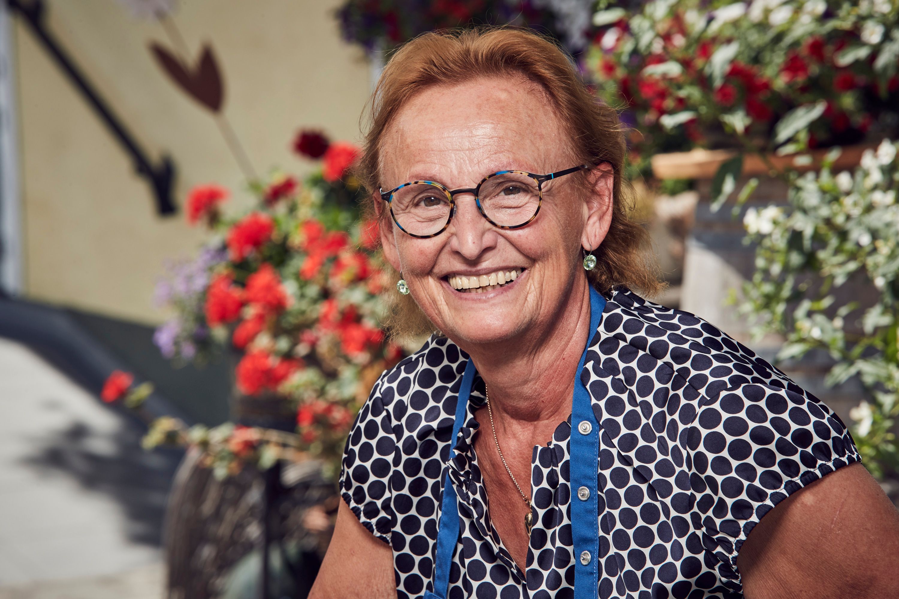 A smiling woman with glasses and a polka-dot blouse in front of flowering plants.