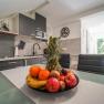 Modern kitchen with fruit bowl on the table, filled with pineapples, apples, oranges and bananas. Dark cupboards and light-colored walls in the background.