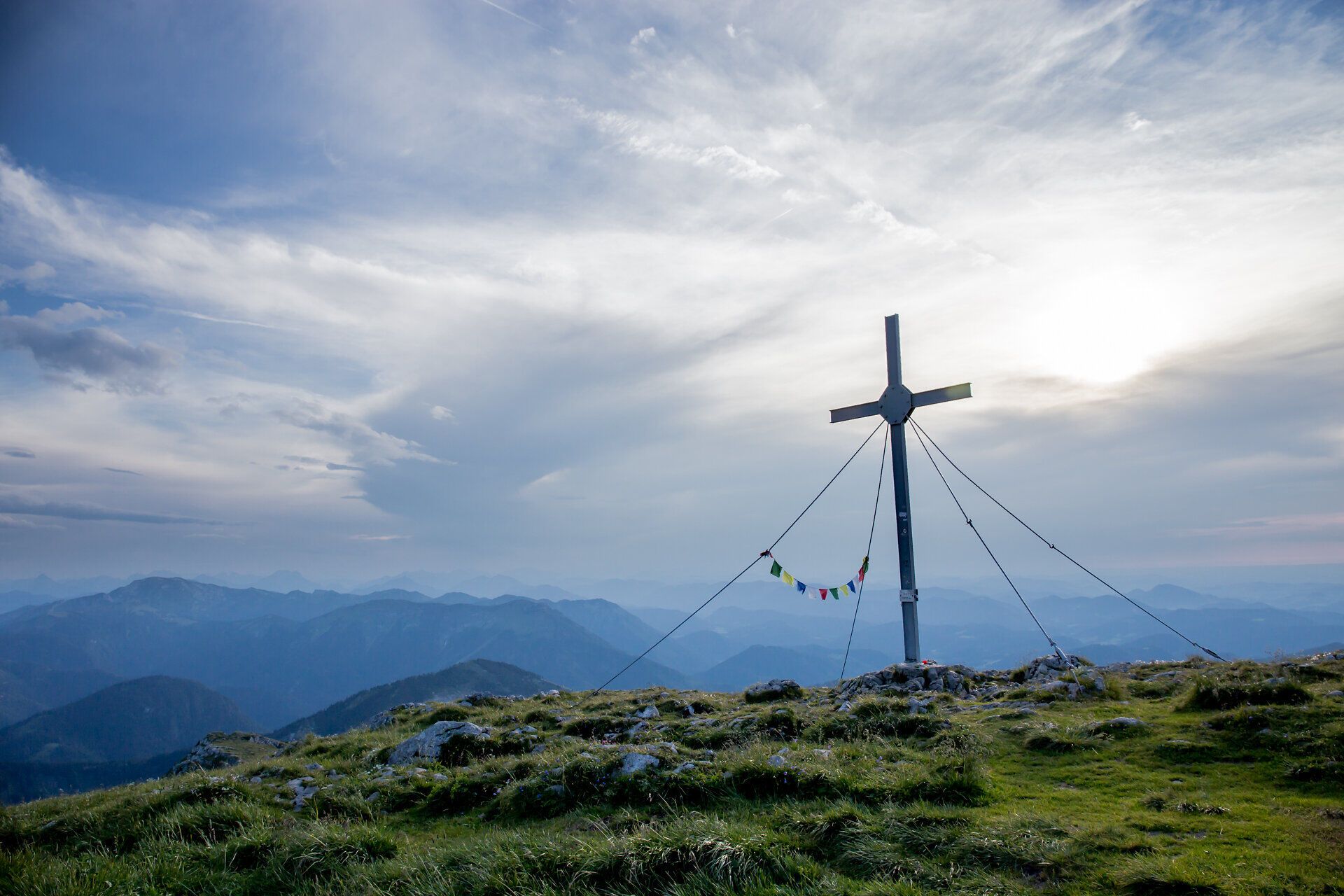 A majestic summit cross towers over the rolling hills, surrounded by a breathtaking mountain landscape. The clouds gently drift across the sky and lend the scenery a peaceful atmosphere, while the fresh mountain air invites you to go hiking.