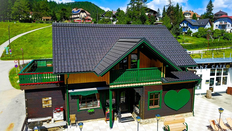 Styrian apartment in summer, © Stefan Zimprich A traditional wooden house with a green heart motif and terrace in summer, surrounded by green hills and blue skies.