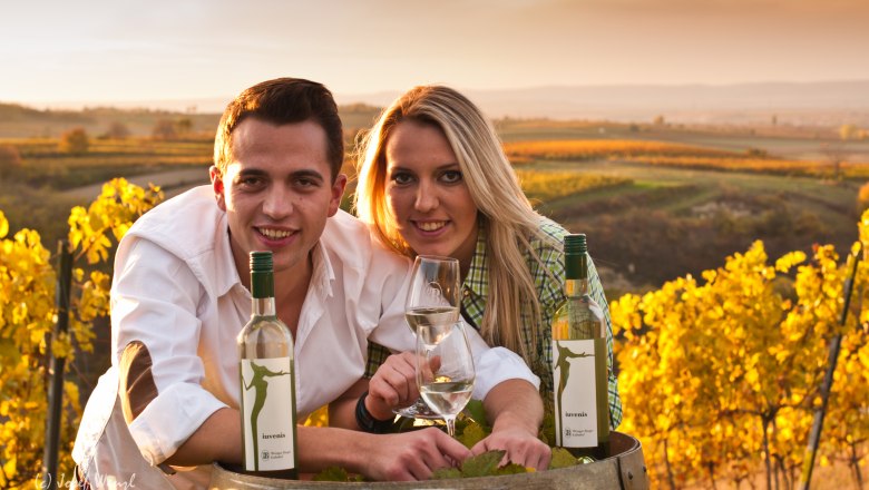 Two people lean over a wine barrel in a vineyard at sunset.