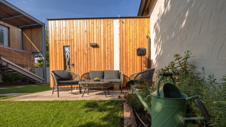 Garden area with seating furniture in front of a wooden house, watering can in the foreground.