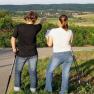 Two women with walking sticks stand on a path and look into the distance, one points in the direction of a village.