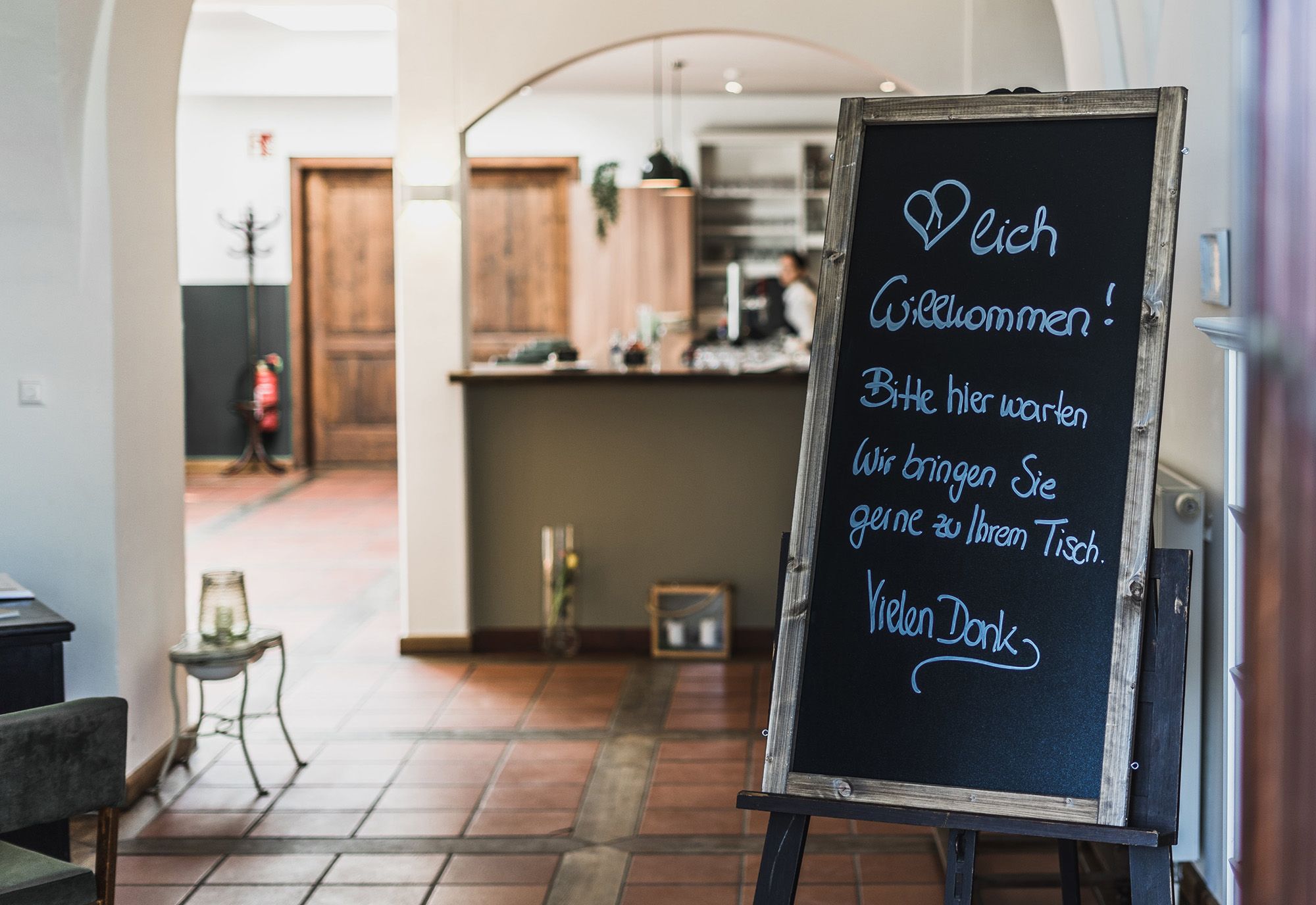 Entrance area of a restaurant with a sign welcoming guests and asking them to wait until they are brought to their table.
