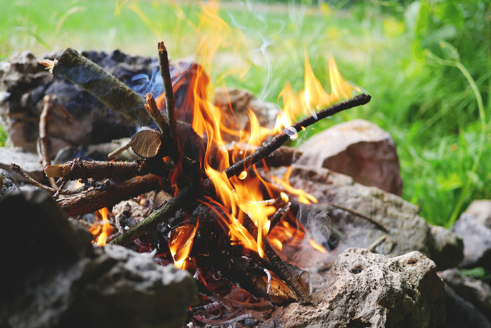 Close-up of a campfire with burning logs and flames, surrounded by stones in the open air.
