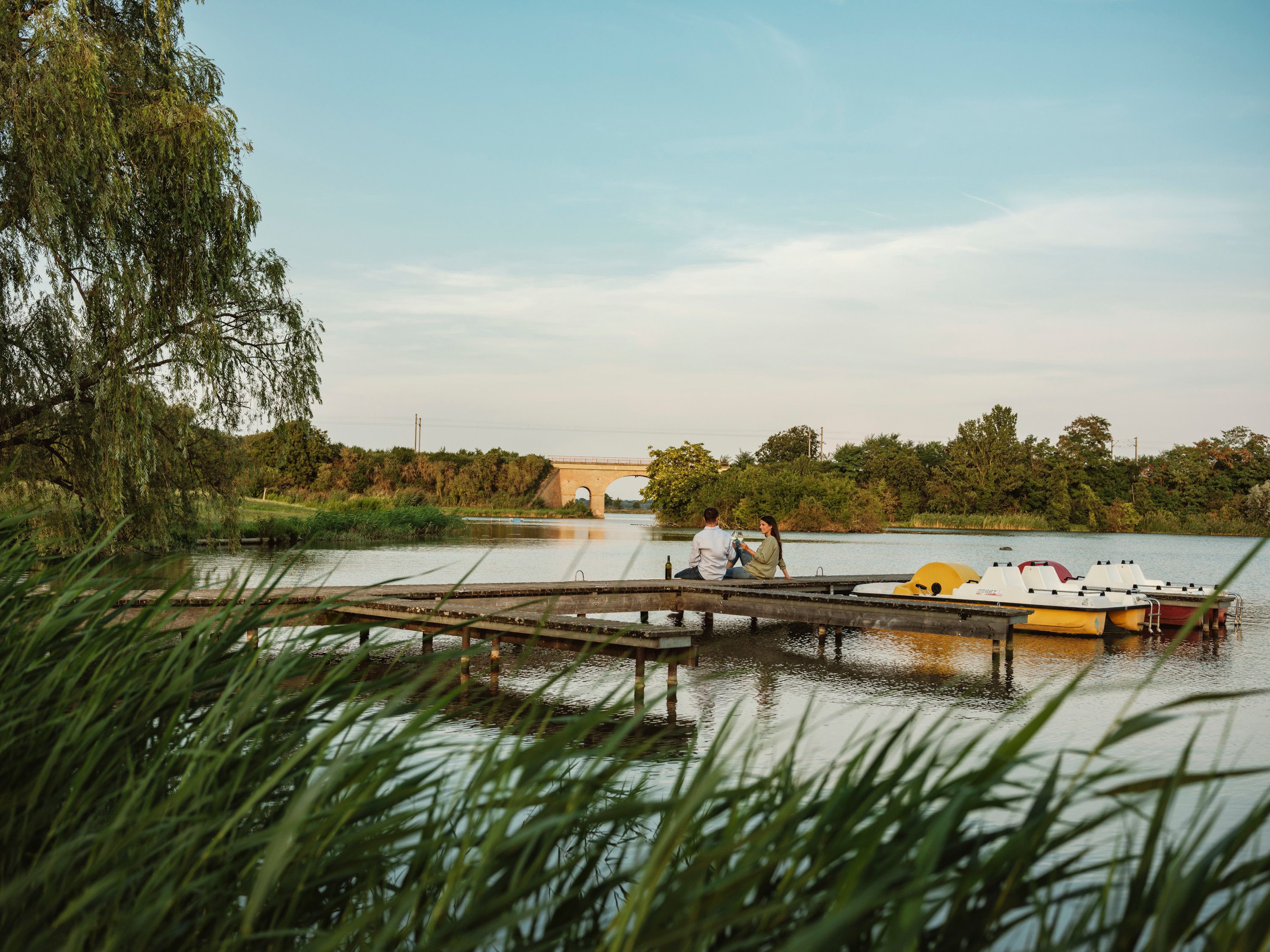 A couple sits on a jetty by a pond in Bernhardsthal, surrounded by nature and pedal boats.