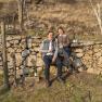 Two people are sitting on a stone wall in a vineyard holding wine glasses.