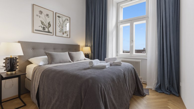 An elegant bedroom with a large bed, gray bed linen, two bedside tables with lamps and a window with a blue curtain.