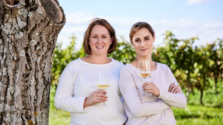 Two women stand with wine glasses in front of a tree in a vineyard.