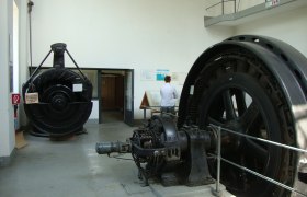 Interior view of a show power plant with large turbines and a person reading an information board.