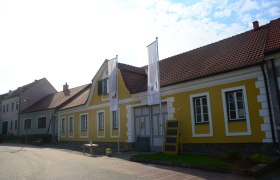 Yellow building with red roof tiles and two flags in front of it.