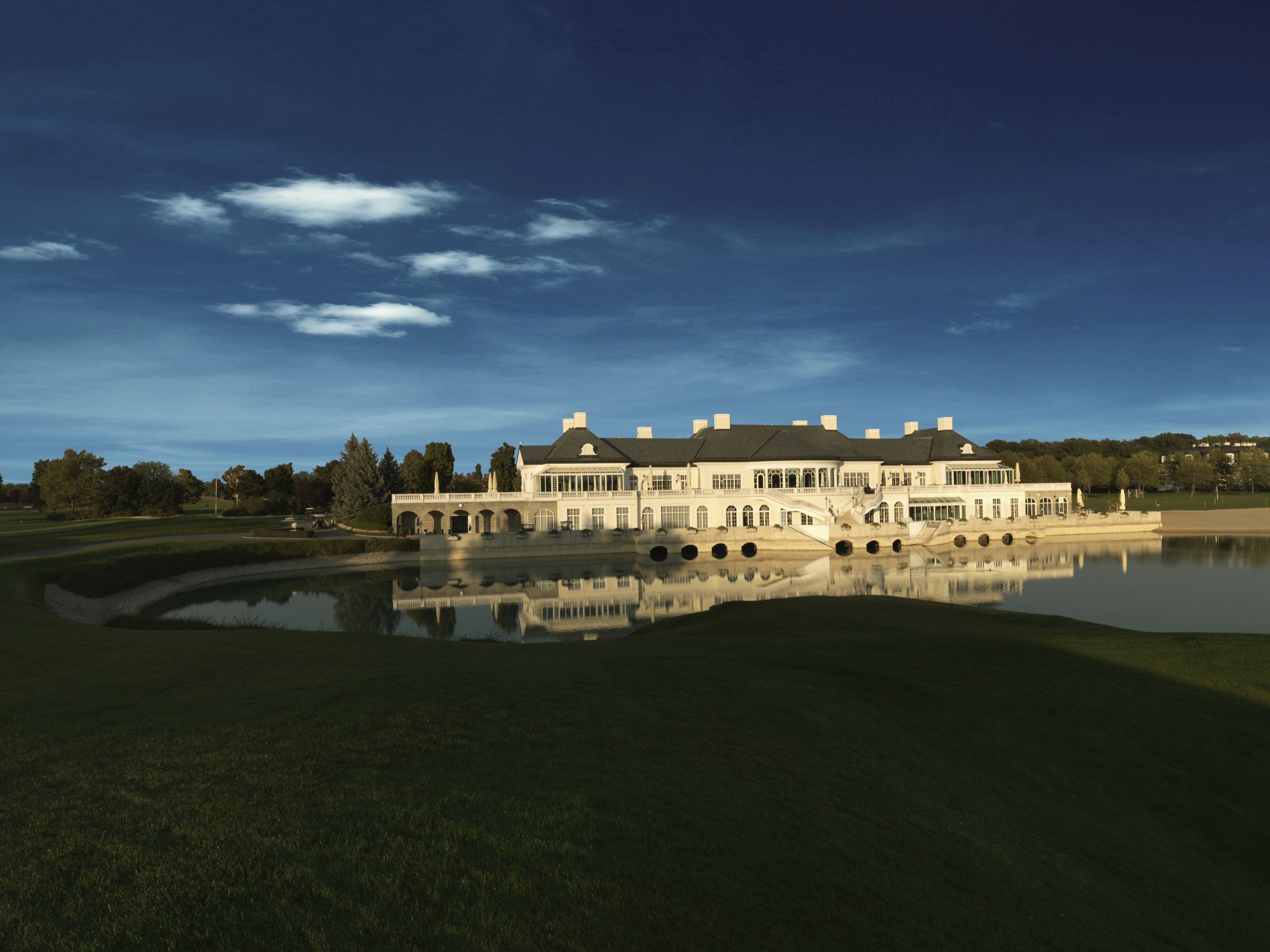 An elegant clubhouse is reflected in a tranquil lake, surrounded by green lawns and trees under a clear blue sky.