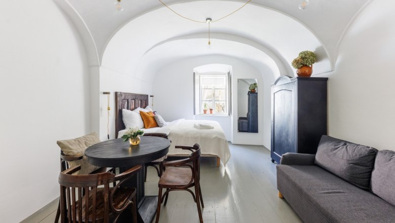 A stylishly furnished guest room with vaulted ceiling, bed, sofa, table and chairs. A window lets in natural light.