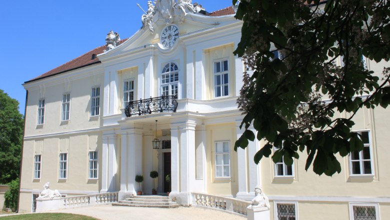 Baroque building with clock and balcony, surrounded by trees.