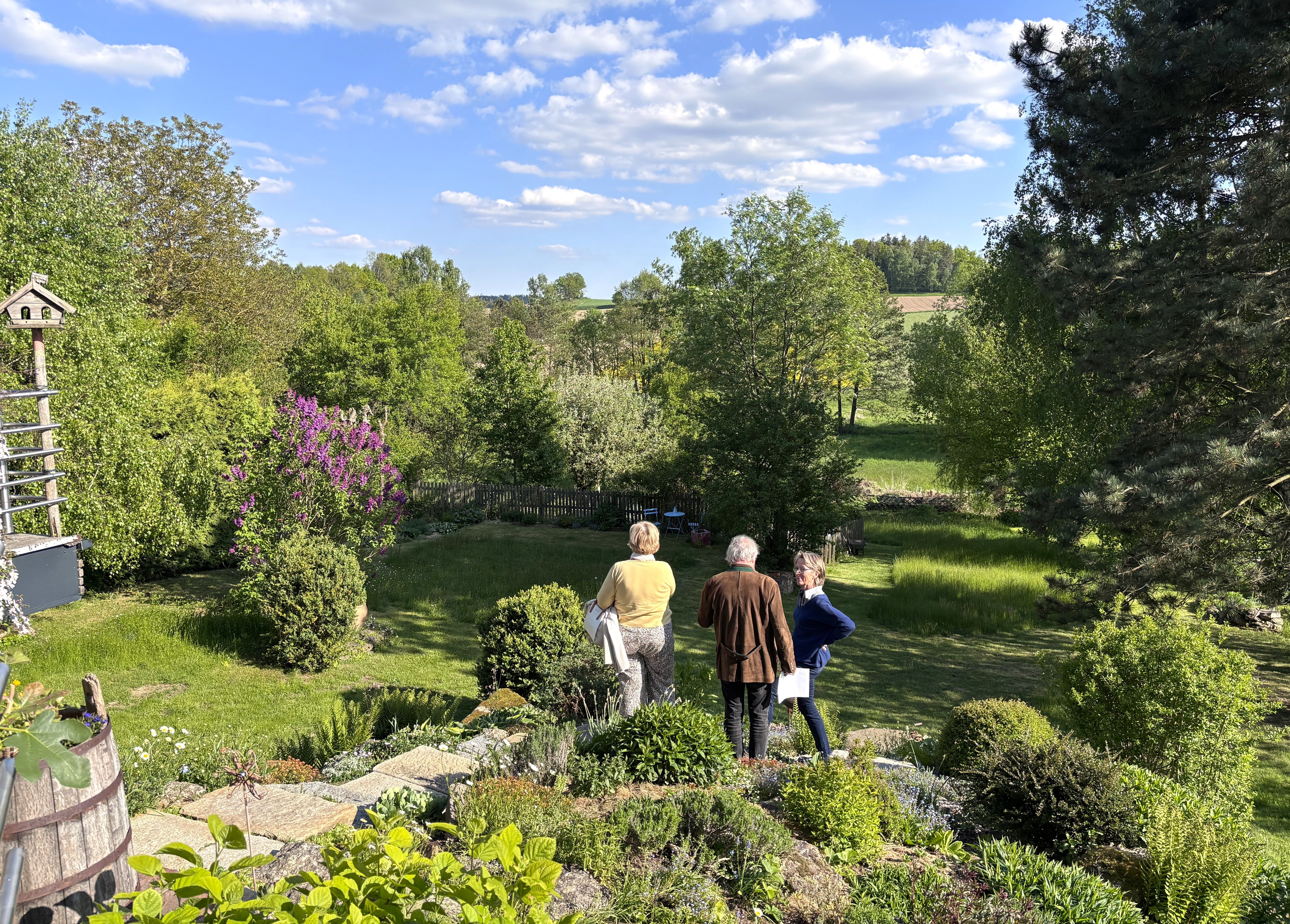 Three people are standing in a green garden with trees and bushes, surrounded by a rural landscape under a blue sky with clouds.