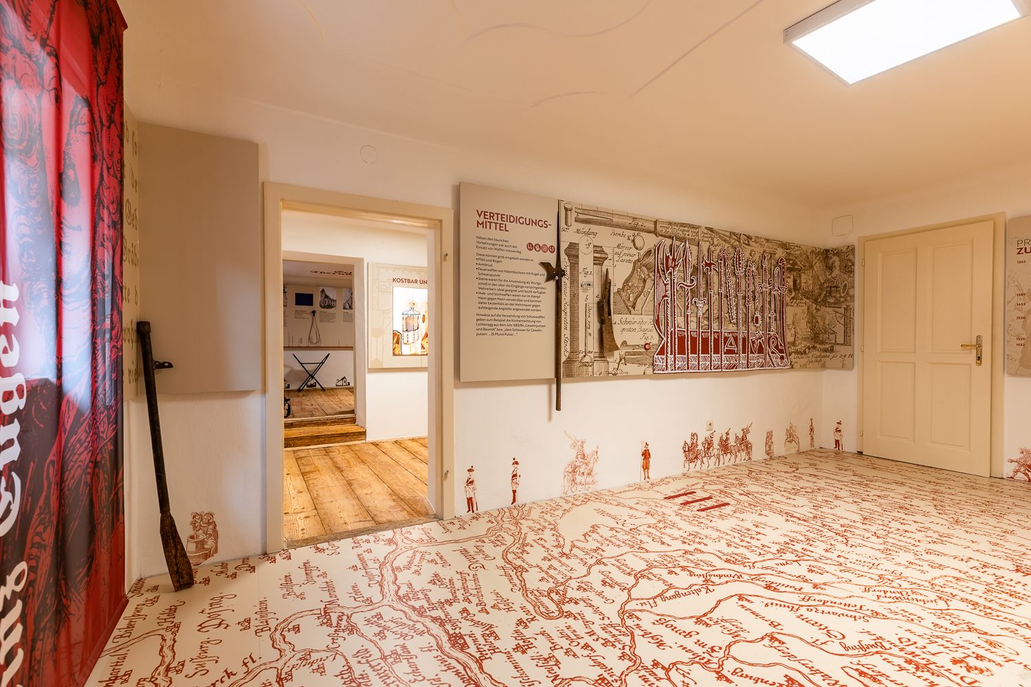 Interior view of a museum room with historical maps and weapons on the walls, a large old map is drawn on the floor