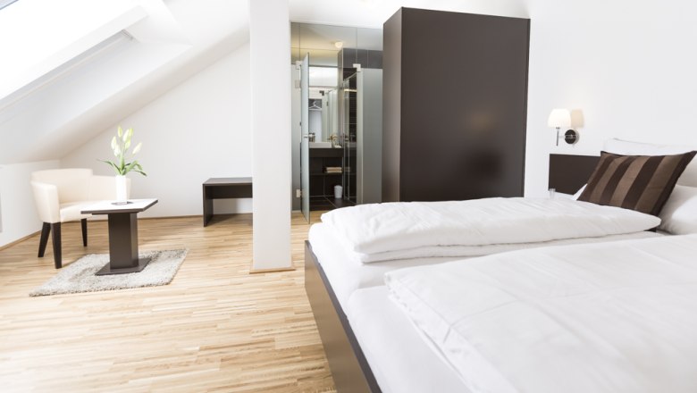 Guest room, © Ernst Jöchlinger Modern guest room with bed, armchair and table under a sloping roof.