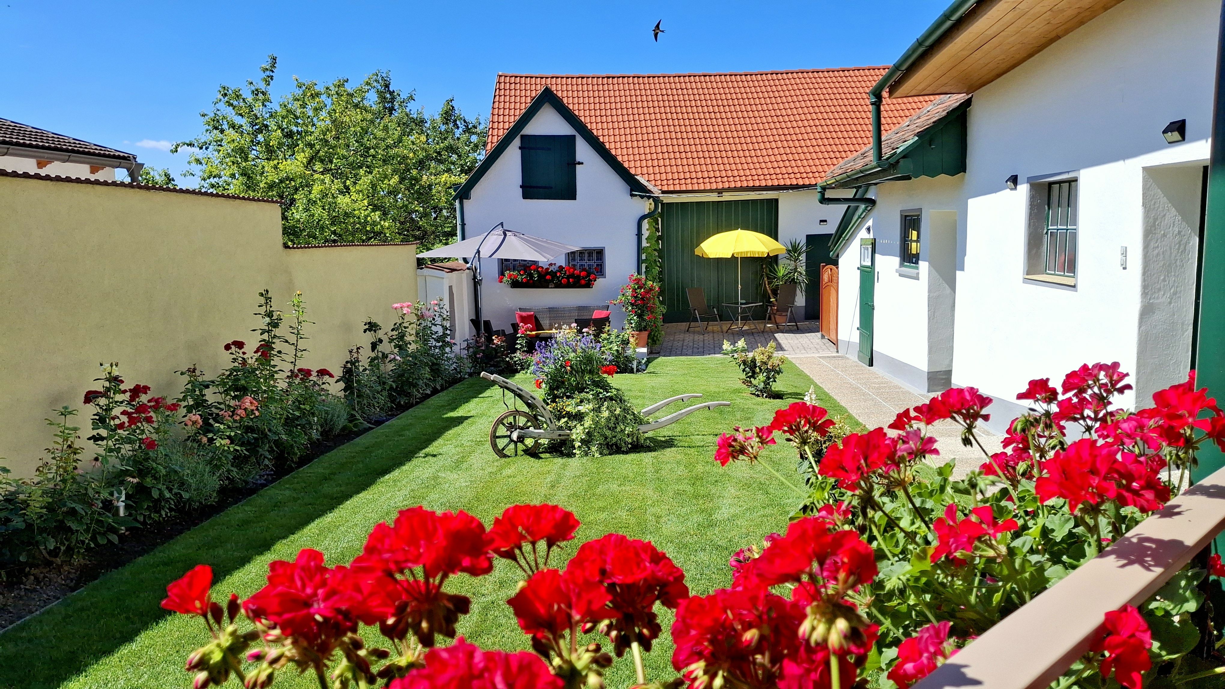 A well-tended garden with red flowers, an old plow and a house with red roof tiles in the background.