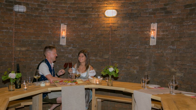 A couple sits at a wooden table in a wine cellar and toasts with red wine.