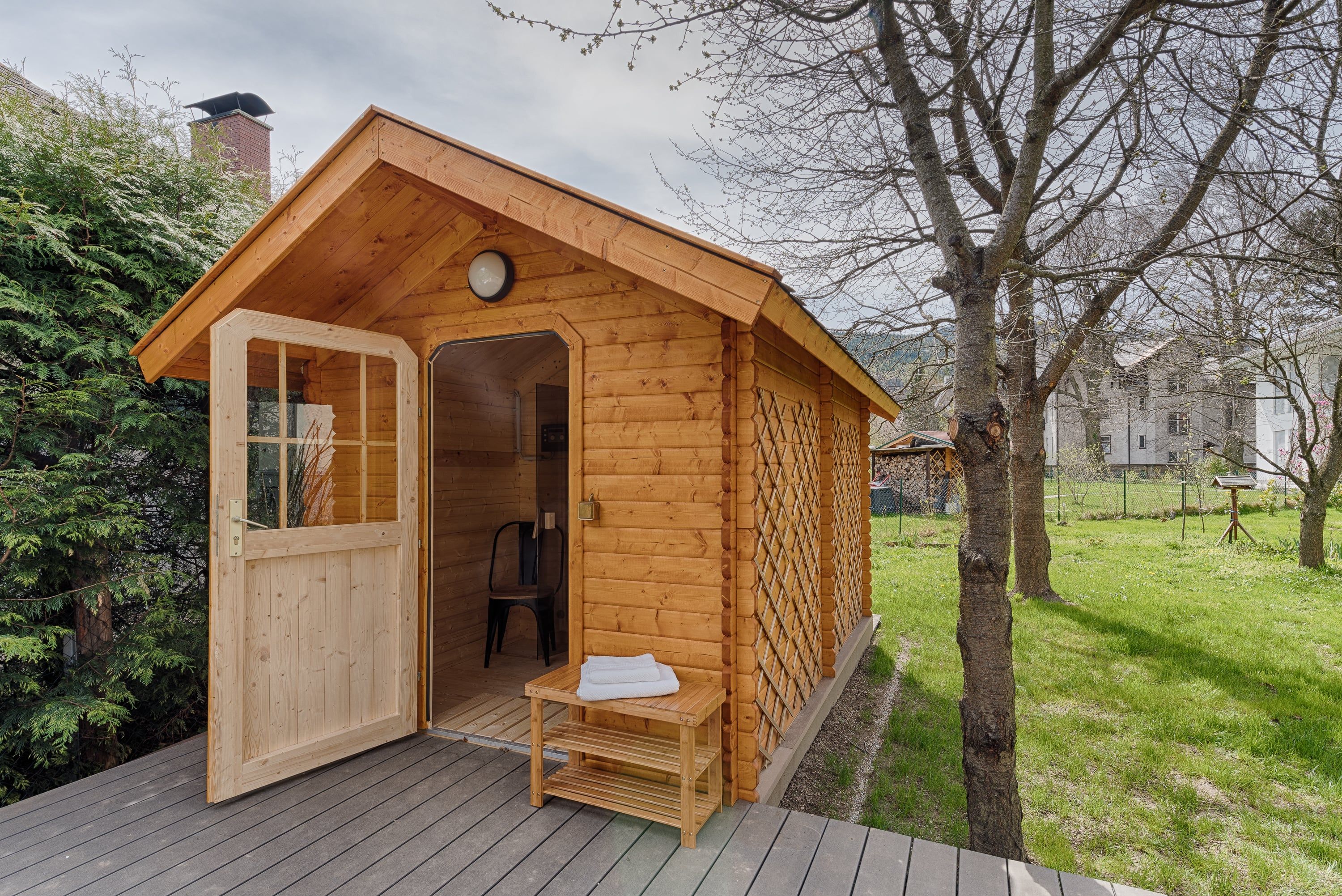 A small wooden sauna in the garden with an open door and towels on a bench in front of it.