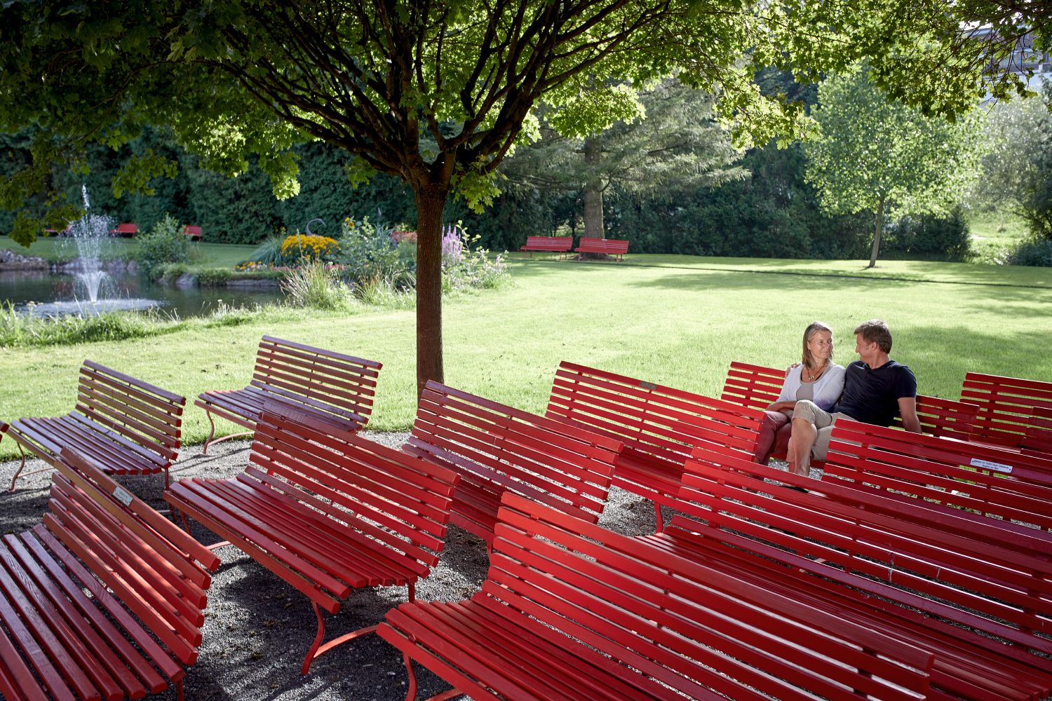 A couple sits on red benches in the spa gardens, surrounded by trees and a pond with a fountain.