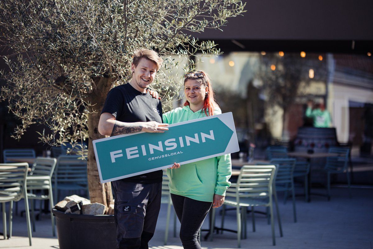 Two people hold a sign reading 'Feinsinn Genusslokal' in front of a tree.
