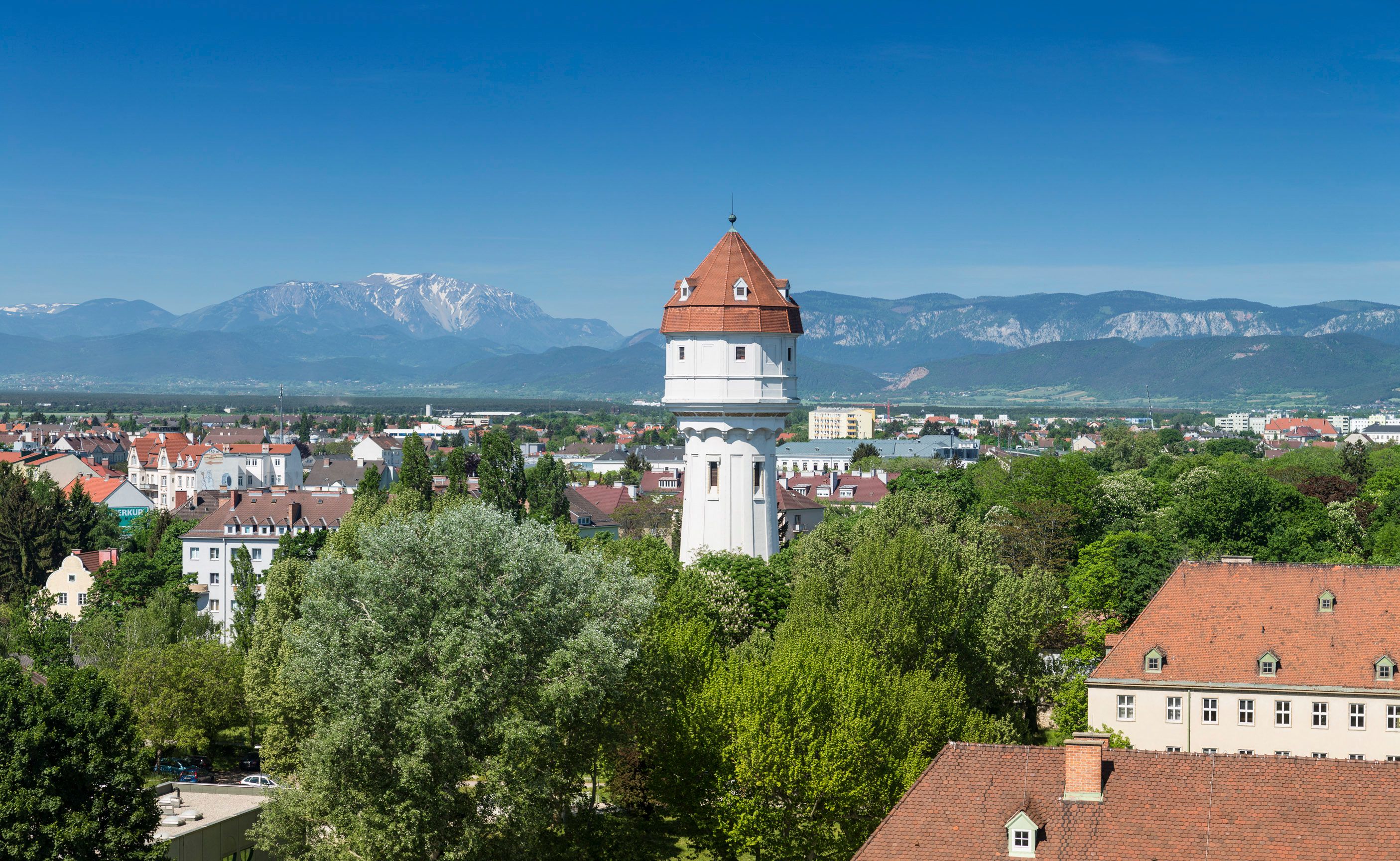 A white water tower with a red roof in a green urban landscape in front of a mountain panorama.