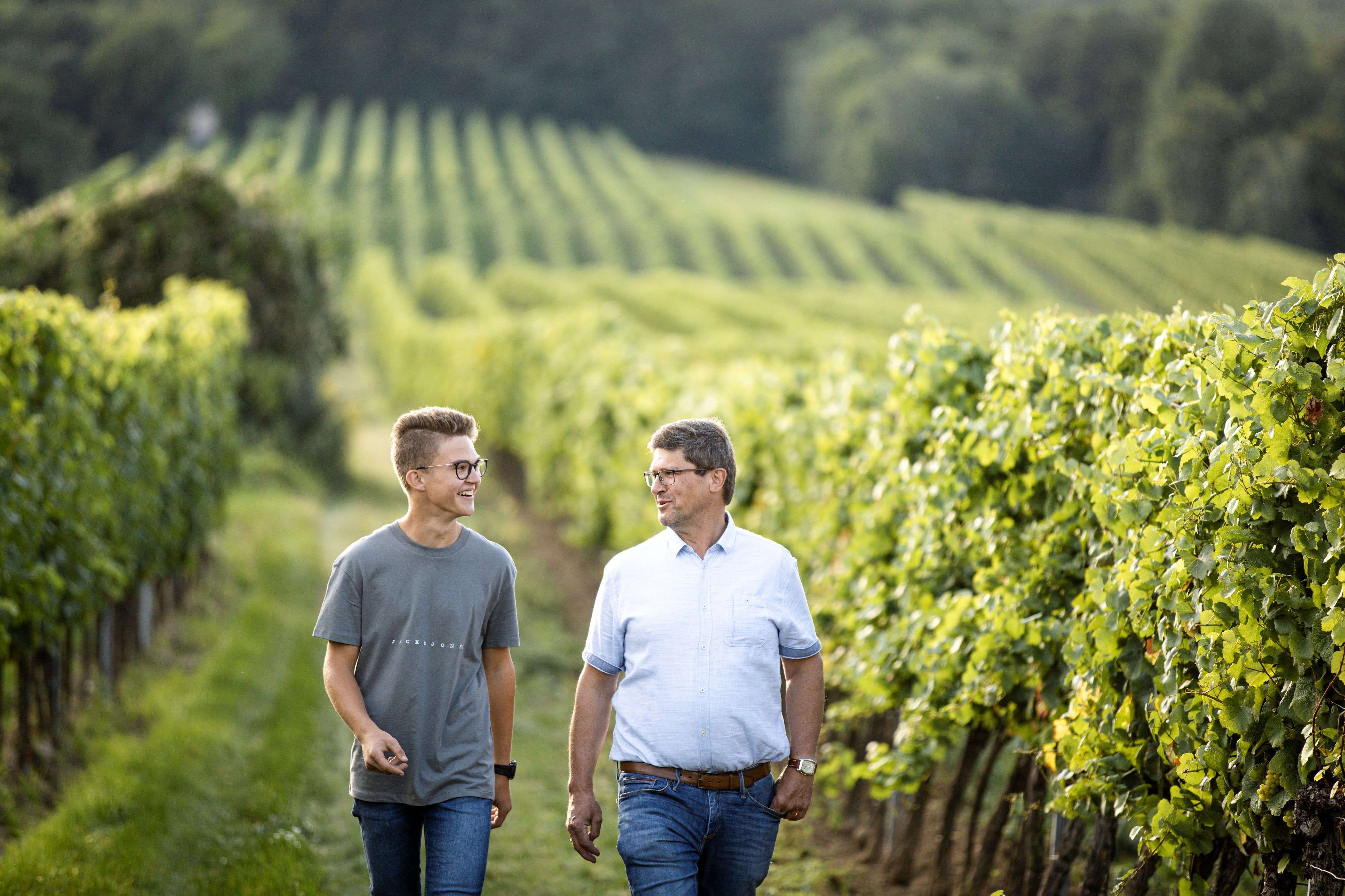 Two men walk through a vineyard smiling.