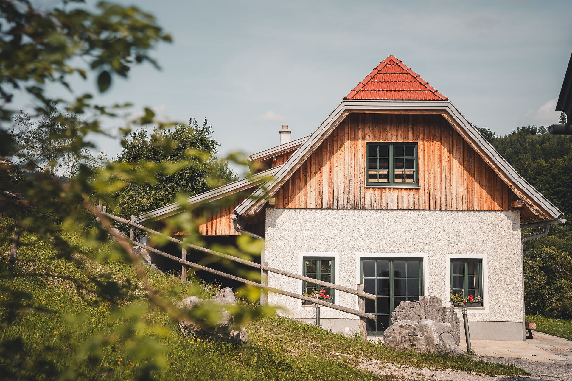 Der Blick fällt auf die Zufahrt zur Rehbergweide, gesäumt von Bäumen und Holzlattenzäunen. Das Bauernhaus wirkt einladend mit offenen Fenstern und Blumenschmuck.