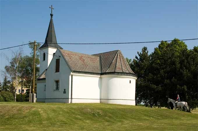 A small white church with a pointed tower stands on a hill. Trees and a rider on a horse can be seen in the background.
