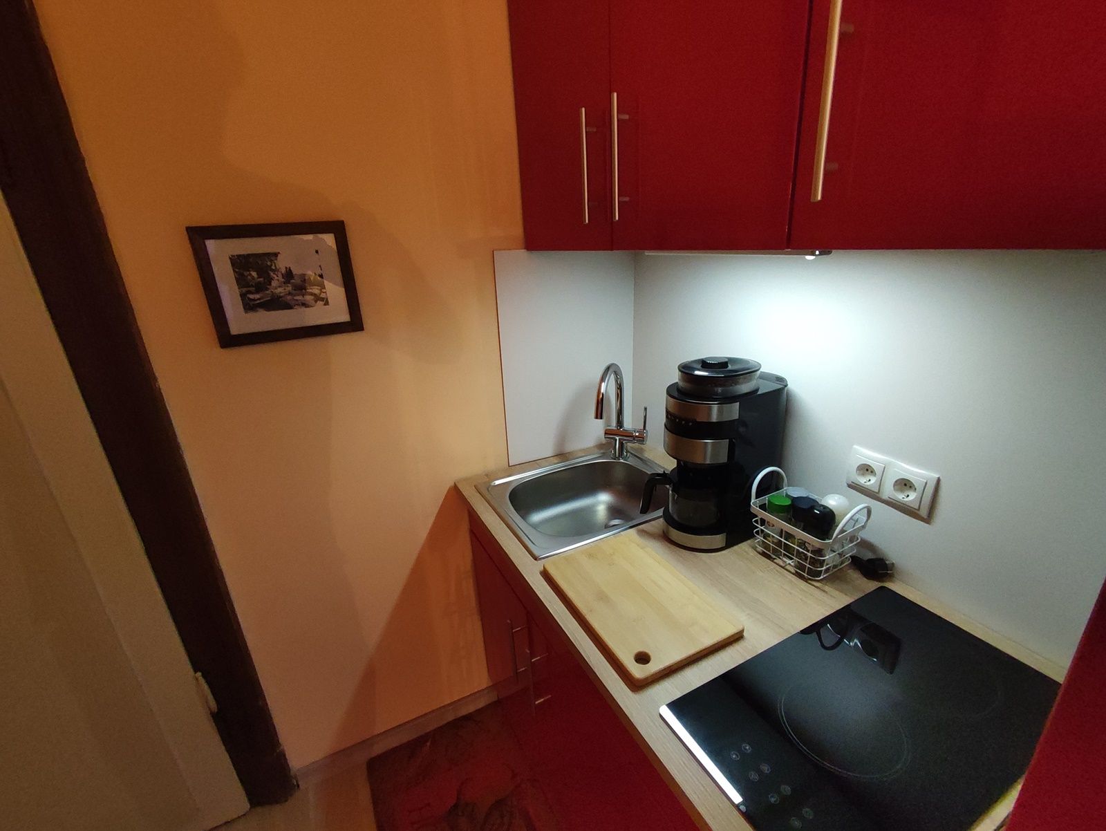 Small kitchen with red cupboard, sink, coffee machine and hob.