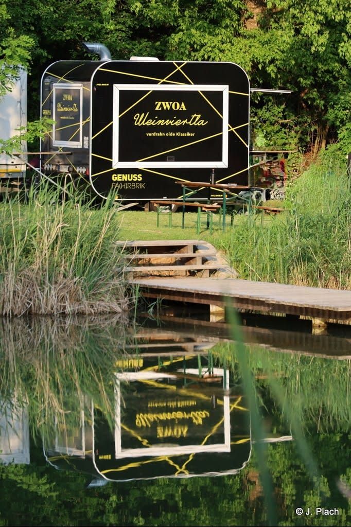 A black food truck with the inscription 'ZWOA Weinviertla' stands in a green setting, reflected in the water.