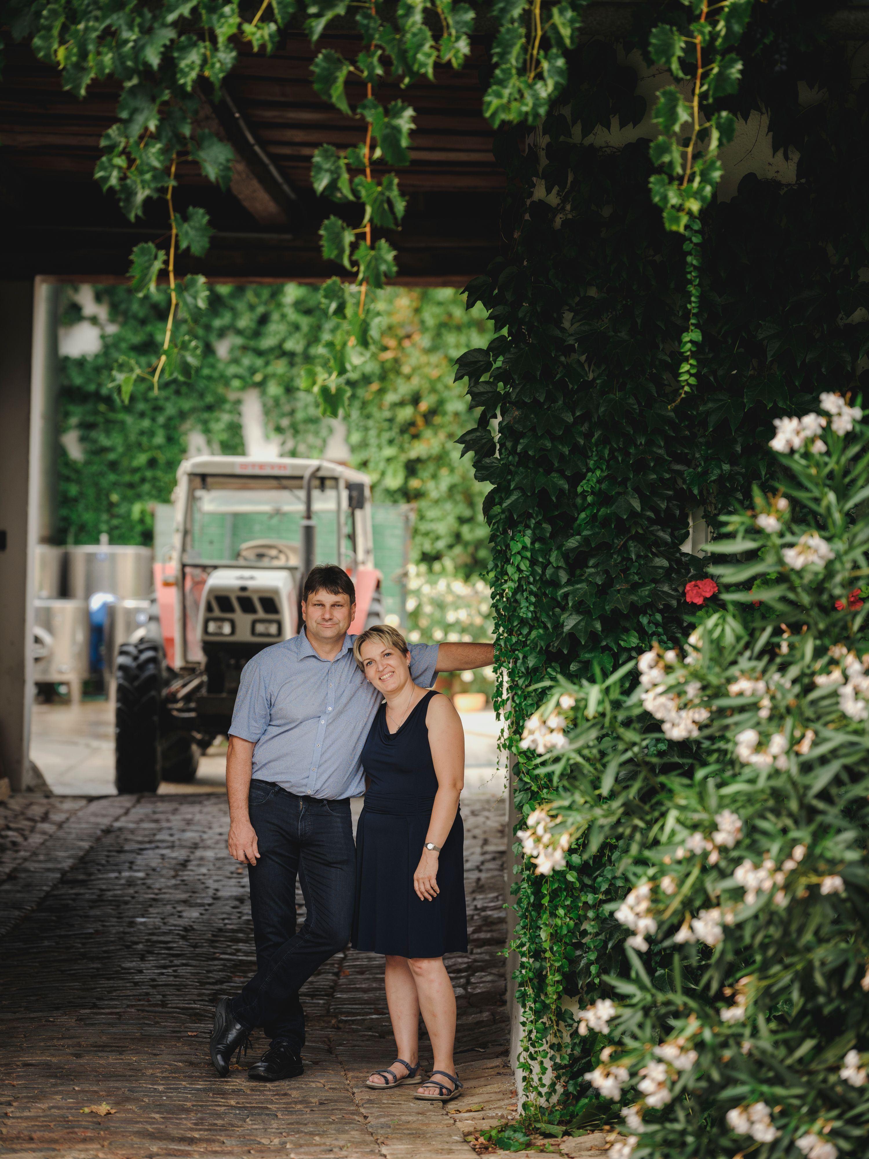 A couple stands under an ivy-covered canopy next to a tractor.