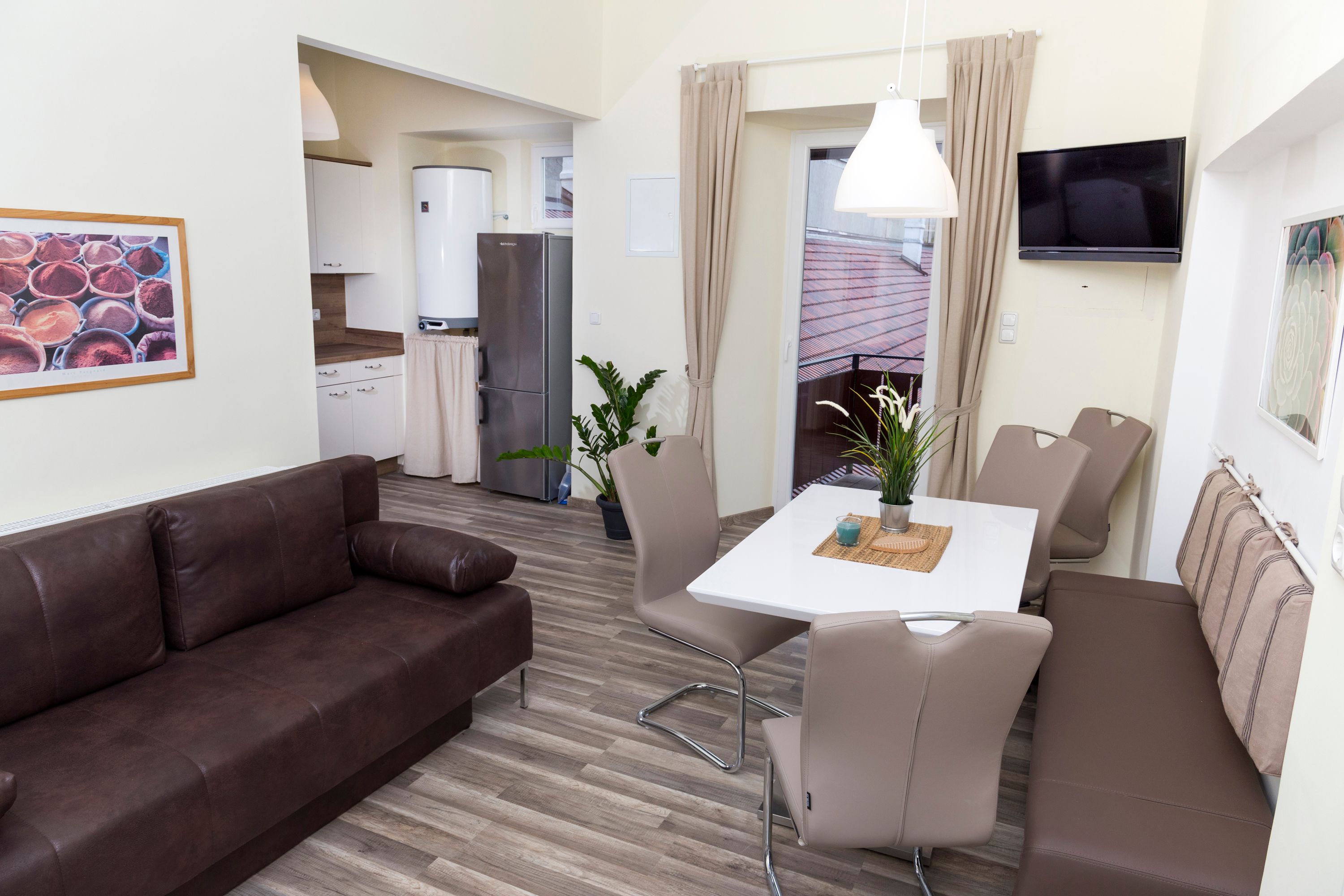 Modern kitchen with brown sofa, dining table and chairs, fridge and plants.