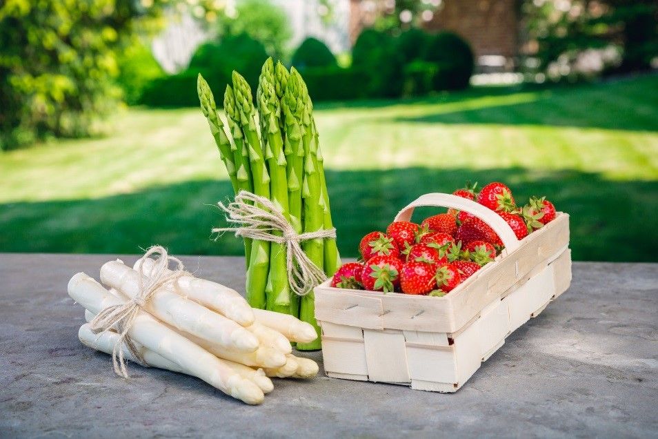 A bunch of green and white asparagus next to a basket of strawberries on an outdoor table.