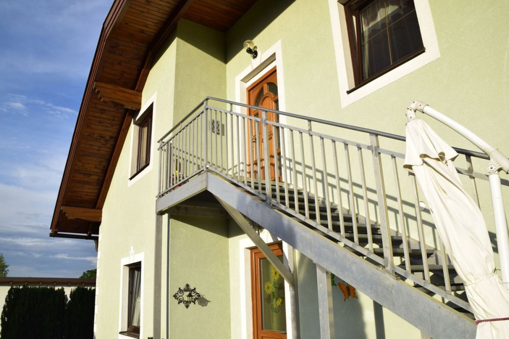 The exterior staircase of a green house with a wooden roof and a parasol.