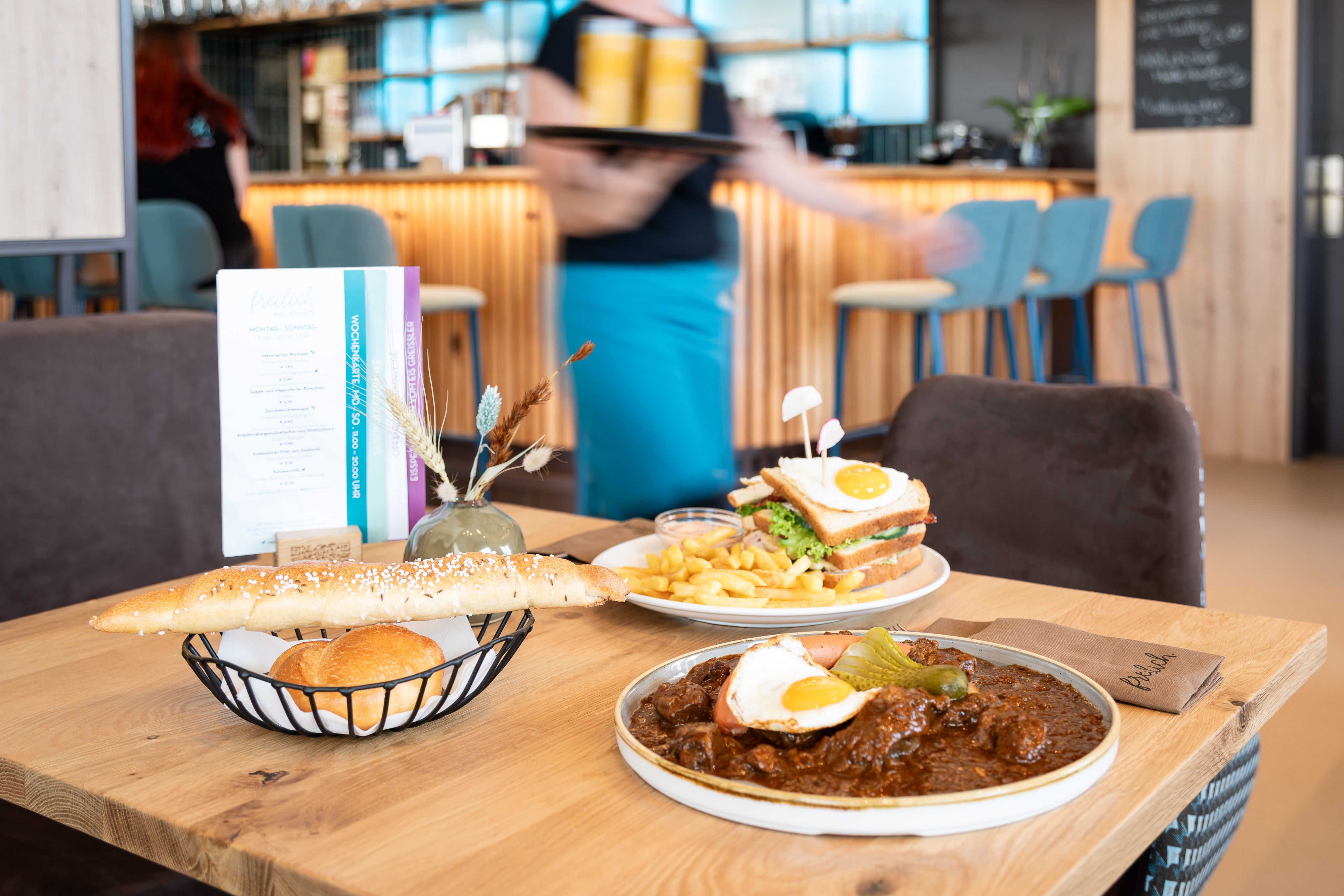 A table in a restaurant with regional dishes: a plate of goulash and fried egg, a sandwich with chips and a bread basket.