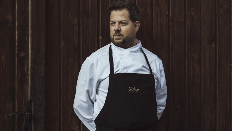 A man in chef's clothes stands in front of a wooden wall.
