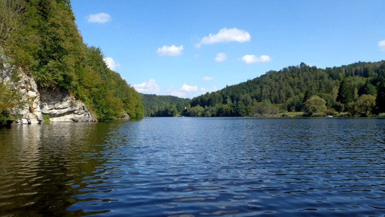 A calm lake surrounded by wooded hills under a blue sky with few clouds.