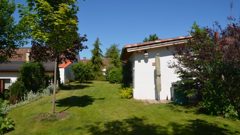 A well-tended garden with lawn, trees and shrubs. Two small buildings with red roofs can be seen in the background.