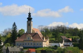 Church with tower in a rural setting, surrounded by trees and houses.
