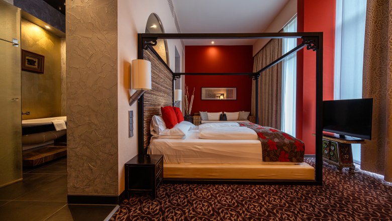 Hotel & Spa Linsberg Asia, © Niederösterreich Werbung / Maximilian Pawlikowsky Elegant hotel room with four-poster bed, red accents and modern bathroom.