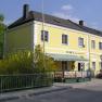 Yellow building with the inscription 'Toni's Bier- und Weinstube', surrounded by green plants and a fence.