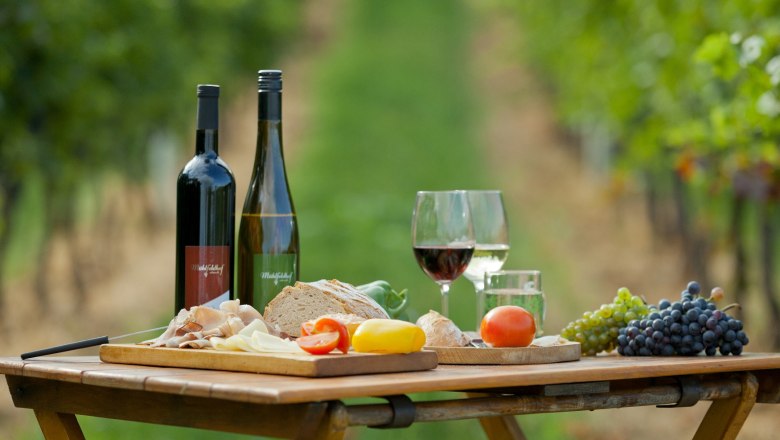 Picnic table with wine bottles, glasses, bread, cheese, ham and grapes in a vineyard.