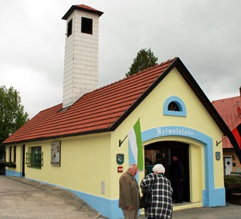 Small yellow building with a red roof and chimney, labeled 'Heimatstube'. Two people are standing in front of it.