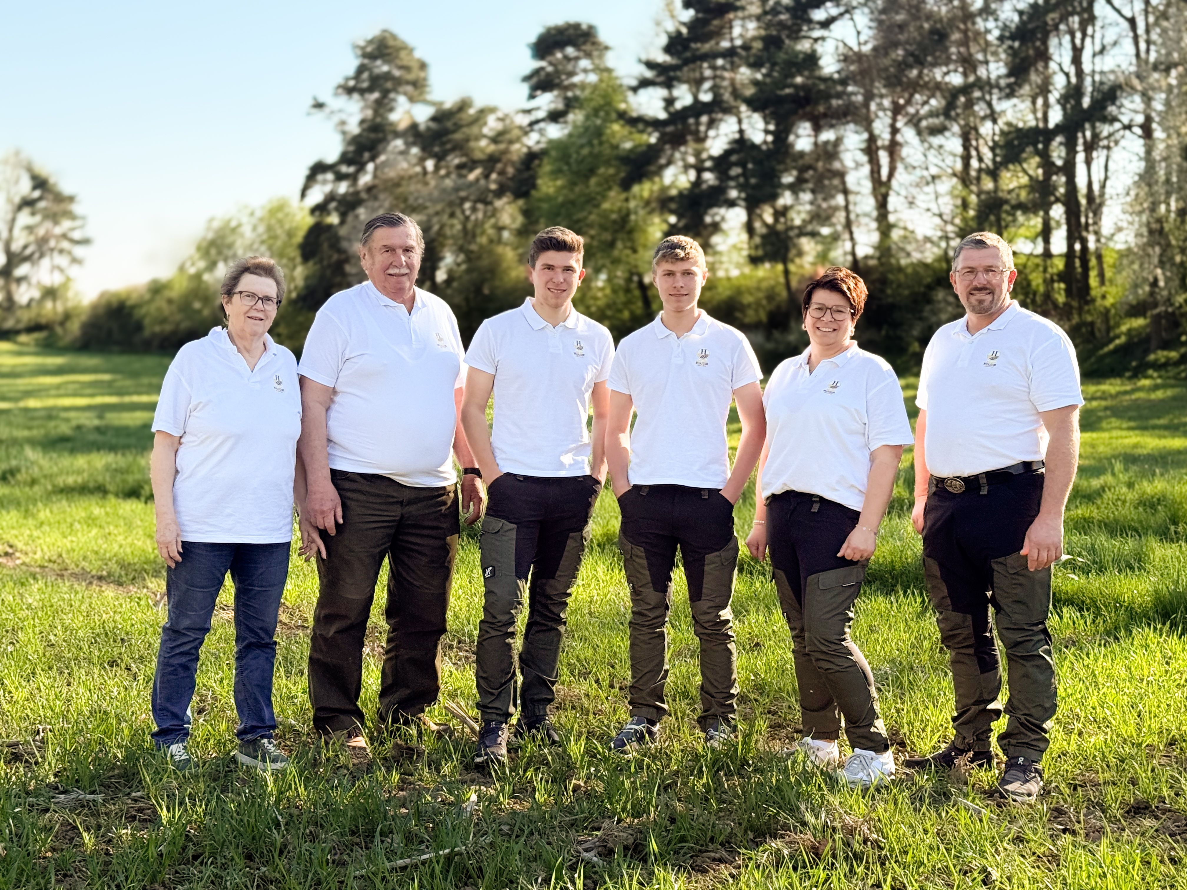 Six people in white polo shirts are standing on a meadow in front of trees.