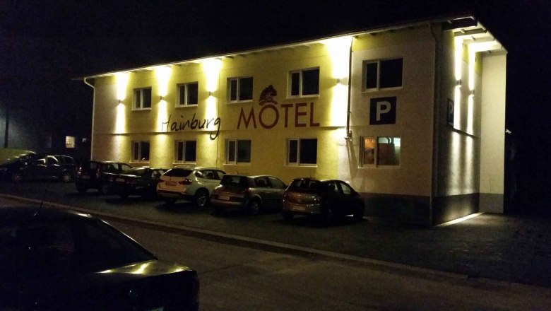 Night shot of an illuminated motel with the lettering 'Hainburg Motel' and parked cars in front of it.